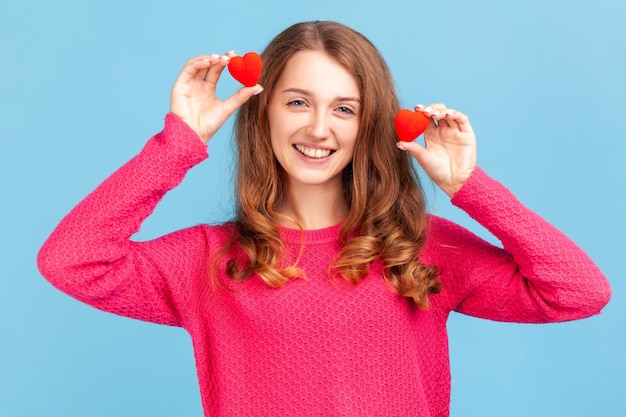 Portrait d'une jeune femme adulte optimiste portant un pull rose, s'amusant avec des coeurs de jouets rouges, montrant son penchant et son dévouement, sa romance. Studio intérieur tourné isolé sur fond bleu.