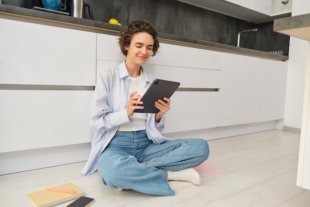 Portrait d'une jeune femme adulte travaillant sur sa tablette assise avec un bloc-notes sur le sol de la cuisine à la maison.