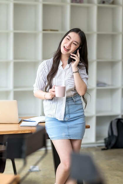 Portrait d'une jeune femme d'affaires asiatique souriante debout tenant une tasse de café et parlant au téléphone portable au bureau