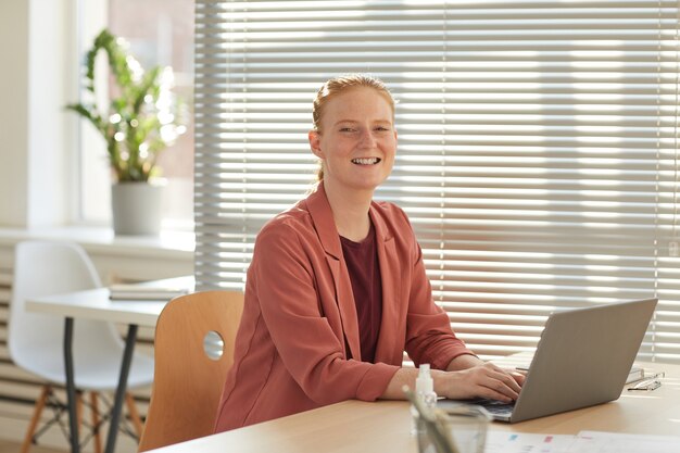 Portrait de jeune femme d'affaires aux cheveux rouges souriant à l'aide d'un ordinateur portable au bureau au bureau ensoleillé