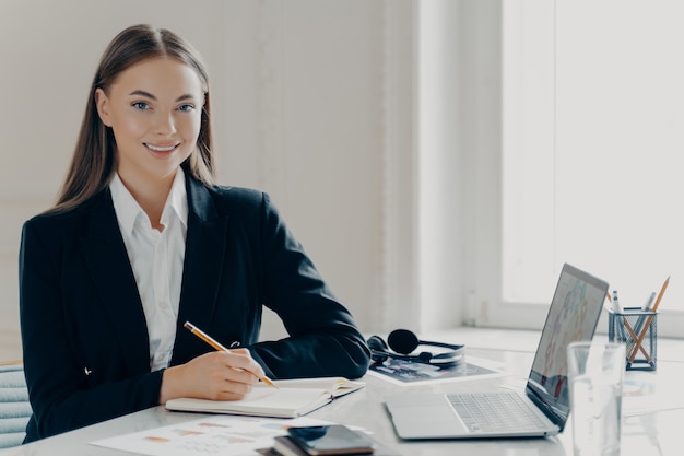 Portrait d'une jeune femme d'affaires caucasienne souriante en costume formel noir regardant la caméra assise près d'un grand bureau blanc avec carnet de notes et ordinateur portable, fenêtre sur fond minimaliste clair.