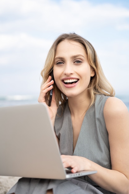 Portrait d'une jeune femme d'affaires joyeuse souriante et parlant au téléphone assise à la plage avec un ordinateur portable