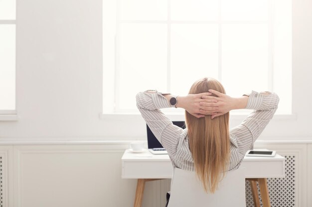 Portrait d'une jeune femme d'affaires mignonne reposant sur une chaise de bureau avec les mains derrière la tête, regardant la fenêtre, espace de copie, vue arrière