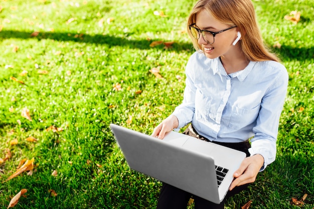 Portrait d'une jeune femme d'affaires portant des lunettes, assis sur l'herbe verte dans le parc, travaillant à l'aide d'un ordinateur portable et d'un casque sans fil