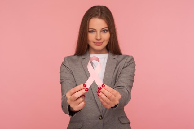 Portrait d'une jeune femme d'affaires positive en veste de costume élégante tenant un ruban rose avertissant du risque de cancer du sein et de la prise de conscience de la société prise en studio sur fond rose