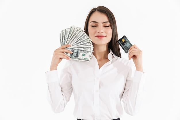 Portrait d'une jeune femme d'affaires séduisante portant des vêtements formels isolés sur un mur blanc, montrant des billets de banque en argent et une carte de crédit