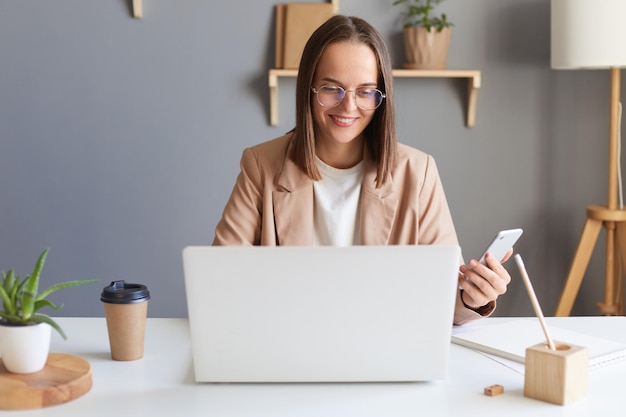 Portrait d'une jeune femme d'affaires souriante et satisfaite travaillant en ligne sur un ordinateur portable dans un bureau à domicile en tapant sur le clavier et en utilisant un téléphone portable pour travailler en regardant l'écran avec un sourire à pleines dents