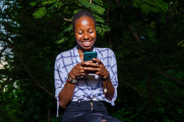 Portrait d'une jeune femme africaine heureuse assise dehors sur un banc lisant un message texte sur son téléphone portable