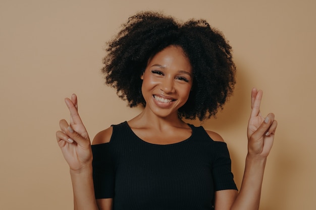 Photo portrait d'une jeune femme africaine heureuse debout isolée sur fond de studio beige foncé avec espace de copie, tenant les doigts croisés pour la bonne chance en souriant largement. émotions positives et langage corporel