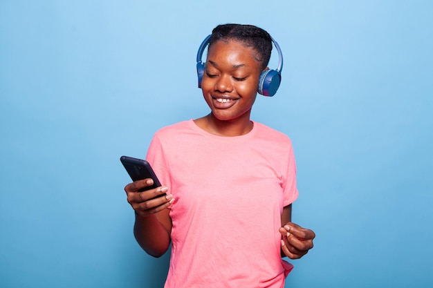 Portrait d'une jeune femme afro-américaine positive portant un casque tenant un smartphone écoutant de la musique debout en studio avec un fond bleu. Adolescent joyeux souriant à la caméra pendant la relaxation