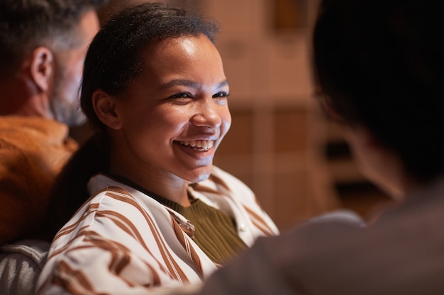 Portrait d'une jeune femme afro-américaine souriant joyeusement en regardant la télévision avec des amis à la maison éclairée par une lumière chaude, espace pour copie