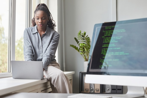 Portrait D'une Jeune Femme Afro-américaine Utilisant Un Ordinateur Portable Assis Près D'une Fenêtre Dans Un Bureau De Développement De Logiciels, écran De Code Au Premier Plan, Espace De Copie