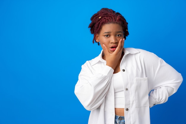 Portrait de jeune femme afro avec l'expression du visage ennuyé touchant son menton sur fond bleu