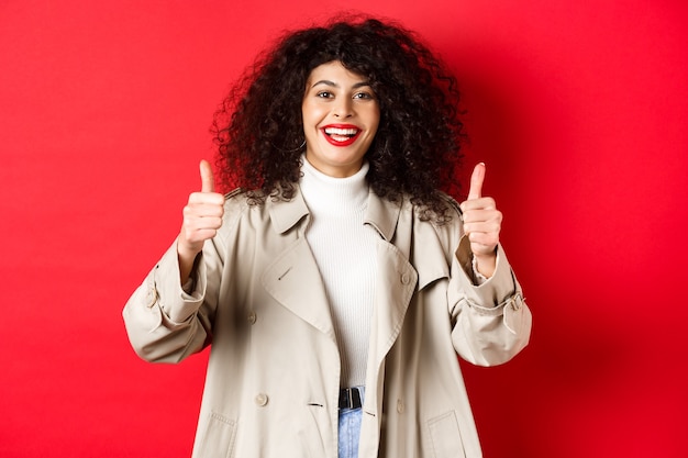 Portrait d'une jeune femme à l'air heureuse portant un trench-coat et montrant le pouce levé dire oui approuver et p...