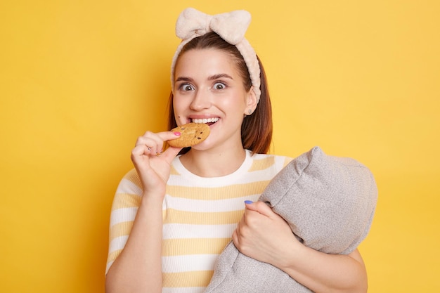 Portrait de jeune femme amusante adulte portant un t-shirt rayé mordant un biscuit mangeant une pâtisserie savoureuse posant isolé sur fond jaune embrassant un oreiller prenant son petit déjeuner