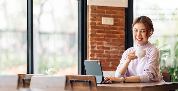 Portrait de jeune femme asiatique à l'aide d'une tablette numérique et se détendre au café en souriant et en regardant la caméra