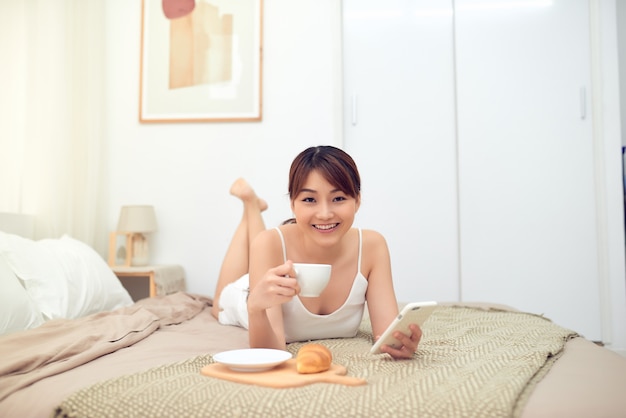 Portrait de jeune femme asiatique appréciant le matin sur le lit tout en utilisant un téléphone portable et en buvant du thé/café