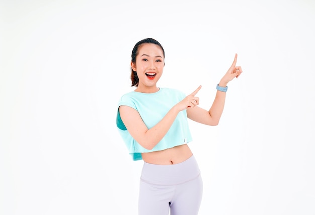 Portrait d'une jeune femme asiatique belle et joyeuse en tenue d'entraînement de gym avec point du doigt sur l'espace de copie isolé sur fond