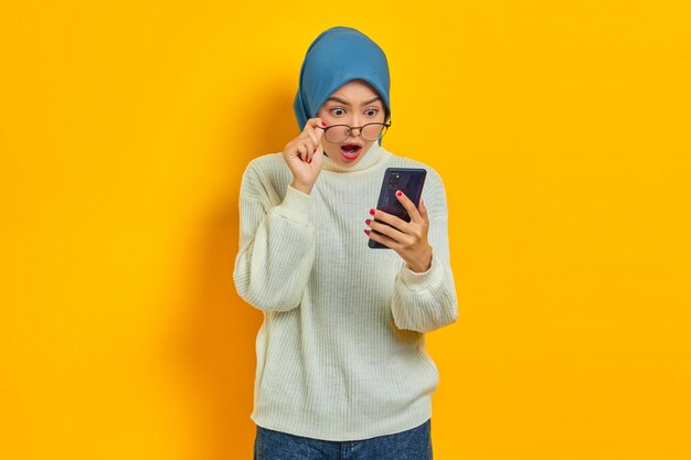 Portrait d'une jeune femme asiatique choquée en chandail blanc et hijab à l'aide d'un téléphone portable prenant des lunettes isolées sur fond jaune Concept religieux de l'islam des gens