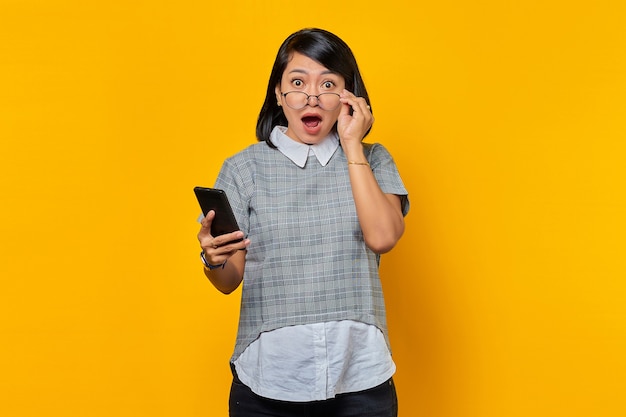 Portrait de jeune femme asiatique choquée et surprise tenant un smartphone et des lunettes regardant la caméra sur fond jaune