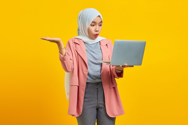 Portrait de jeune femme asiatique choquée tenant un ordinateur portable et pointant isolé sur fond jaune