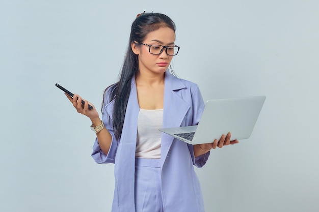 Portrait de jeune femme asiatique debout à l'aide d'un ordinateur portable et tenant un téléphone portable avec une expression pensive isolé sur fond blanc