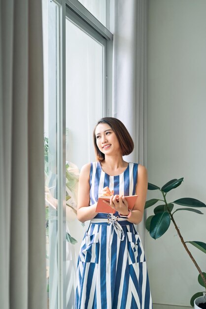 Portrait de jeune femme asiatique debout écrivant dans un cahier regardant par la fenêtre