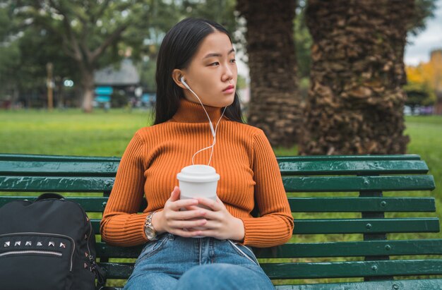 Portrait de jeune femme asiatique écoutant de la musique avec des écouteurs et tenant une tasse de café dans le parc à l'extérieur.