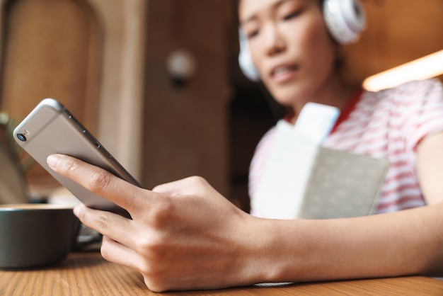 Portrait de jeune femme asiatique écoutant de la musique avec des écouteurs et tenant un téléphone portable dans un café à l'intérieur