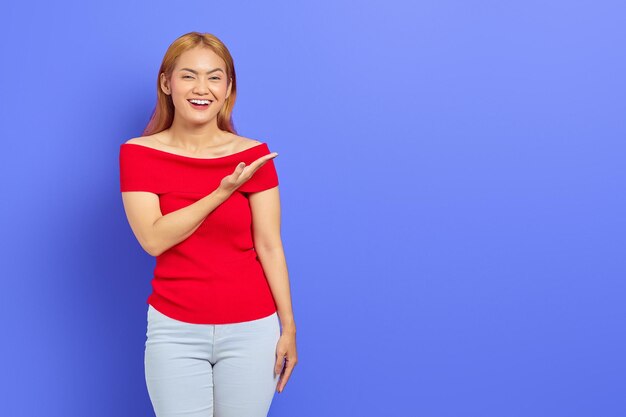 Portrait d'une jeune femme asiatique excitée en robe rouge debout et présentant un espace de copie avec des palmiers isolés sur fond violet