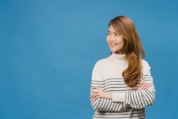 Portrait d'une jeune femme asiatique avec une expression positive, les bras croisés, un large sourire, vêtue de vêtements décontractés et regardant l'espace sur le mur bleu