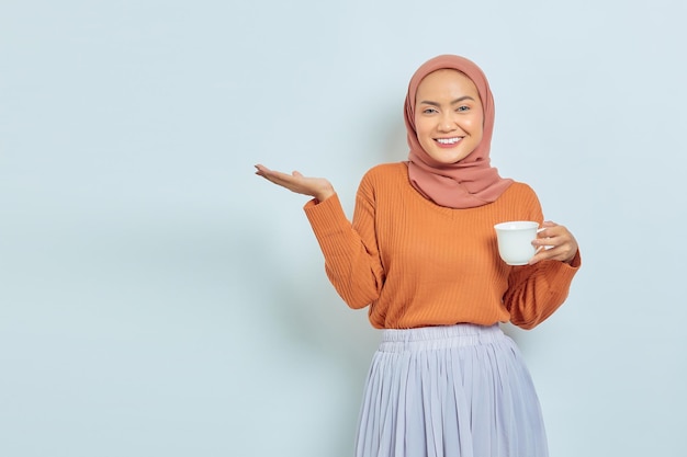 Portrait d'une jeune femme asiatique gaie en pull marron tenant une tasse et pointant vers l'espace de copie avec des palmiers isolés sur fond blanc