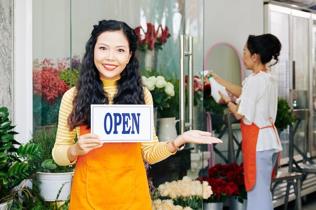 Portrait d'une jeune femme asiatique heureuse tenant une pancarte ouverte lors de l'invitation d'un client dans son magasin de fleurs