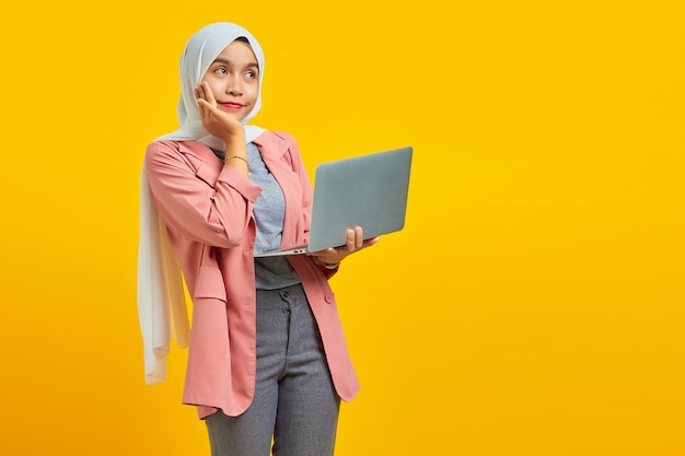 Portrait d'une jeune femme asiatique joyeuse tenant un ordinateur portable et pensant à quelque chose d'isolé sur fond jaune