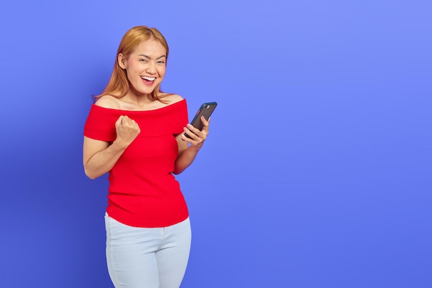 Portrait d'une jeune femme asiatique joyeuse tenant un smartphone tout en montrant un geste gagnant isolé sur fond violet
