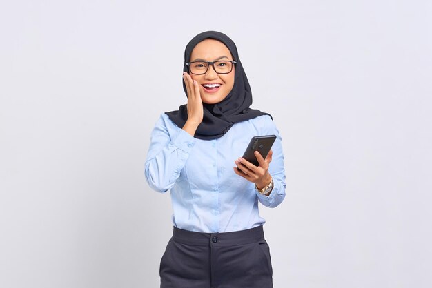 Portrait d'une jeune femme asiatique joyeuse tenant un téléphone portable et lisant de bonnes nouvelles isolées sur fond blanc