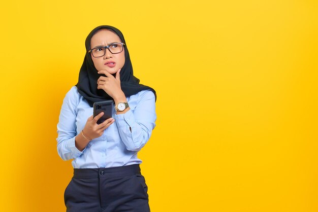 Portrait de jeune femme asiatique pensive à l'aide d'un téléphone mobile et levant isolé sur fond jaune