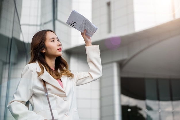 Portrait d'une jeune femme asiatique portant une chemise blanche tout en couvrant le soleil par un journal dans les rues du centre-ville. Elle sort du bâtiment après les travaux de finition.