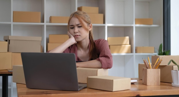 Un portrait d'une jeune femme asiatique sérieuse travaillant avec un ordinateur portable dans le bureau rempli de colis et de boîtes empilant une table à la recherche occupée pour la livraison des PME concept de démarrage d'entreprise et de bureau à domicile