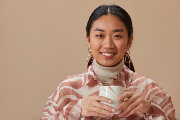 Portrait de jeune femme asiatique souriant à l'avant tenant une tasse avec du thé isolé sur un mur beige