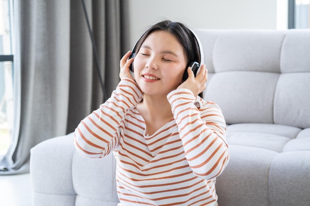 portrait d'une jeune femme asiatique souriante aux yeux fermés écoutant de la musique dans un casque sans fil à la maison