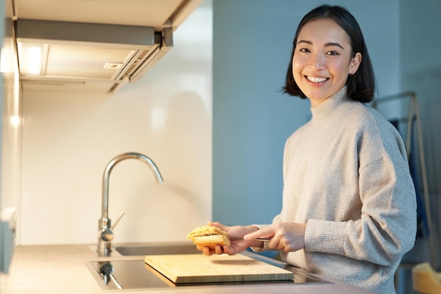 Portrait d'une jeune femme asiatique souriante debout dans la cuisine et faisant un sandwich cuisinant pour elle-même