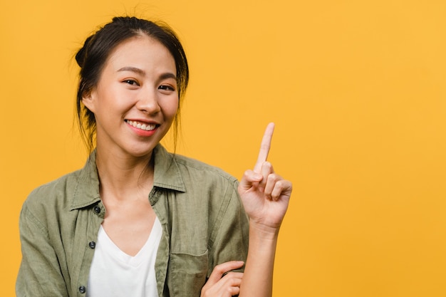 Portrait d'une jeune femme asiatique souriante avec une expression joyeuse, montre quelque chose d'étonnant dans un espace vide dans un tissu décontracté isolé sur un mur jaune. Concept d'expression faciale.