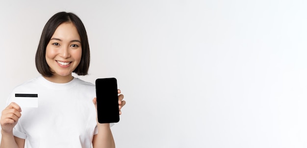 Portrait d'une jeune femme asiatique souriante montrant un écran de téléphone portable et une carte de crédit debout sur fond blanc