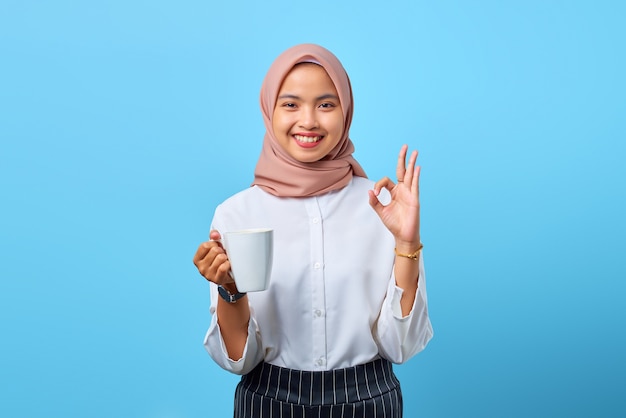 Portrait d'une jeune femme asiatique souriante montrant un signe d'accord et tenant une tasse sur fond bleu