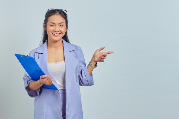 Portrait d'une jeune femme asiatique souriante tenant un dossier de documents et pointant du doigt l'espace de copie isolé sur fond violet