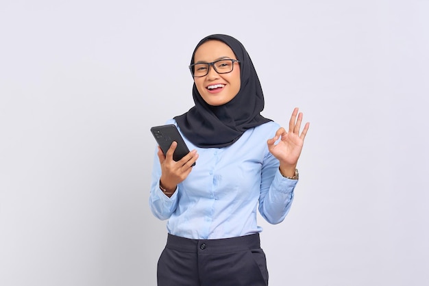 Portrait de jeune femme asiatique souriante tenant un téléphone portable et gesticulant signe d'accord isolé sur fond blanc