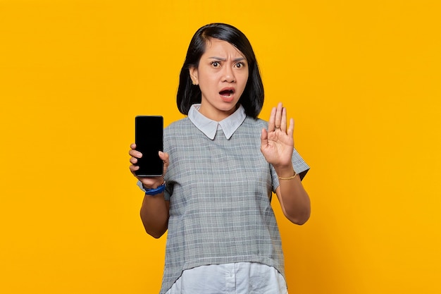 Portrait d'une jeune femme asiatique surprise avec la bouche ouverte montrant un smartphone à écran blanc et faisant un geste d'arrêt avec les paumes