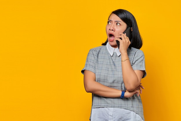 Portrait d'une jeune femme asiatique surprise et choquée lors de la réception d'un appel entrant sur un smartphone sur fond jaune