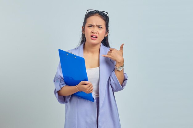 Portrait d'une jeune femme asiatique surprise en costume tenant un dossier de documents et faisant mon geste d'appel isolé sur fond violet